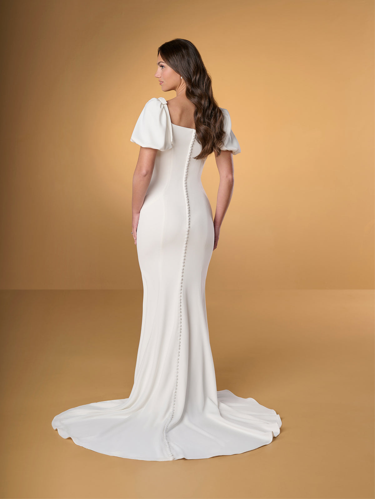 Woman wearing a white wedding dress with a plain background / Slim crepe dress with square neckline and puffy short sleeves. Buttons at the back along zipper closure and all the way down on train.