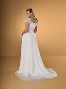 A woman modeling a white wedding gown from the back against a warm tan studio backdrop. The dress has short puff sleeves, a fitted corset-style back, and a full flowing skirt with a slight train. / A-line gown in crinkled organza and satin midriff area on bodice only. Short puffy sleeves. Covered buttons along zipper closure at the back dress.