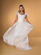 A woman modeling a white off-the-shoulder ballgown with a fitted bodice and a voluminous flowing skirt, posed against a warm tan studio backdrop. / A-line gown in crinkled organza and satin midriff area on bodice only. Short puffy sleeves. Covered buttons along zipper closure at the back dress.