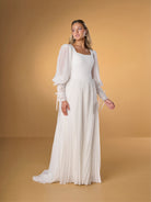 A woman modeling a long white maxi dress with sheer balloon sleeves, a square neckline, and a pleated skirt, posed against a warm tan studio backdrop. / A-line style in chiffon with square neckline shape, permanently pleated chiffon on skirt area only. Puffy sleeves fitted on long cuffs, decorated with narrow ribbons.