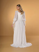 A woman modeling a long white dress with sheer balloon sleeves, tie details at the cuffs, and a pleated skirt, posed against a warm tan studio backdrop. / A-line style in chiffon with square neckline shape, permanently pleated chiffon on skirt area only. Puffy sleeves fitted on long cuffs, decorated with narrow ribbons.