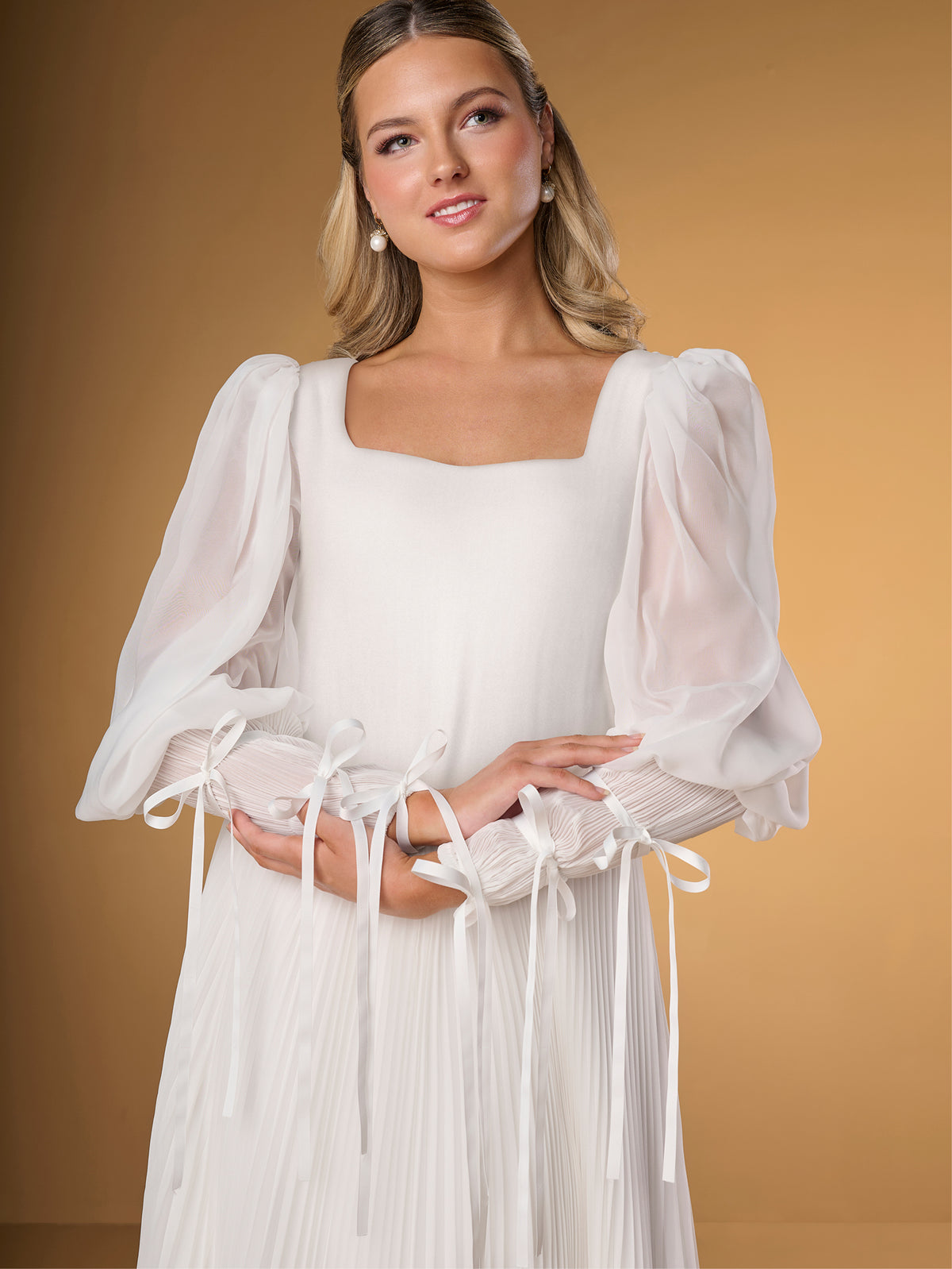 Woman wearing a white dress with sheer sleeves against a beige background / A-line style in chiffon with square neckline shape, permanently pleated chiffon on skirt area only. Puffy sleeves fitted on long cuffs, decorated with narrow ribbons.
