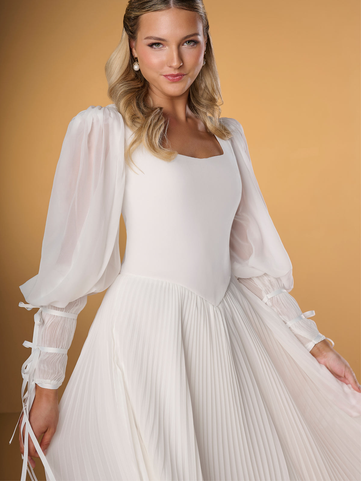 A woman modeling a white wedding dress with a structured bodice, square neckline, sheer balloon sleeves with lace-up cuffs, and a pleated skirt, posed against a warm tan studio backdrop. / A-line style in chiffon with square neckline shape, permanently pleated chiffon on skirt area only. Puffy sleeves fitted on long cuffs, decorated with narrow ribbons.