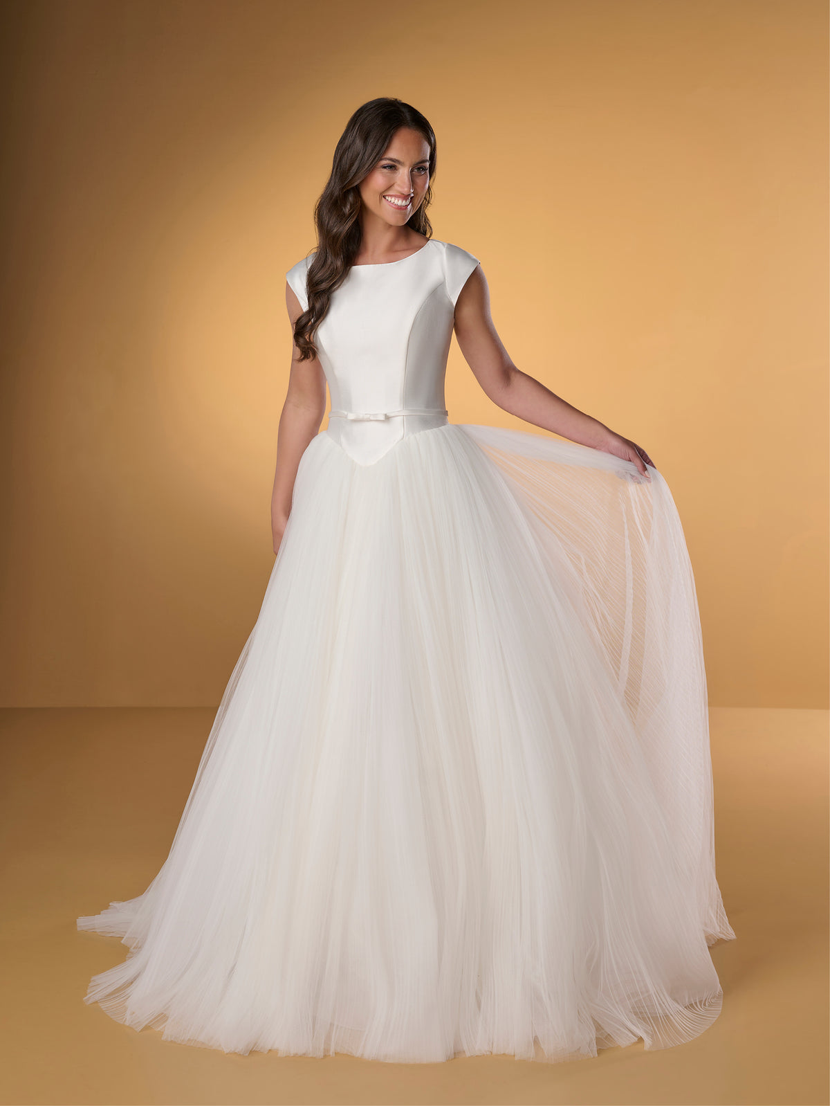 Woman in a white wedding dress on a yellow background / Ball gown with Mikado bodice and pleated tulle skirt. Bateau neckline shape, cup sleeves, narrow detachable waist band with little bow on center front. Covered buttons along zipper closure at the back.