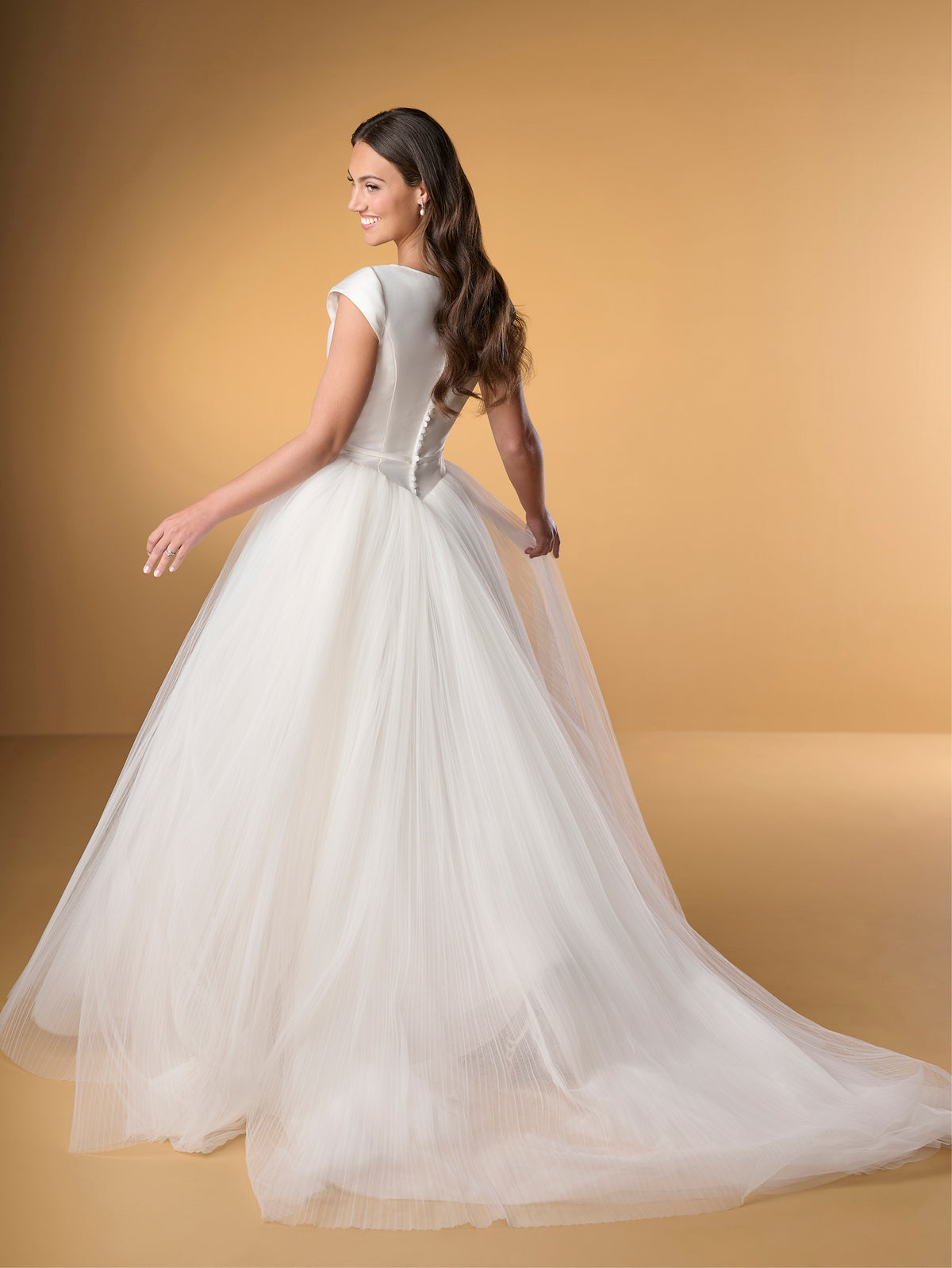 Woman in a white wedding dress on a beige background / Ball gown with Mikado bodice and pleated tulle skirt. Bateau neckline shape, cup sleeves, narrow detachable waist band with little bow on center front. Covered buttons along zipper closure at the back.