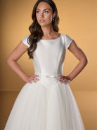 Woman wearing a white dress against a gold background / Ball gown with Mikado bodice and pleated tulle skirt. Bateau neckline shape, cup sleeves, narrow detachable waist band with little bow on center front. Covered buttons along zipper closure at the back.