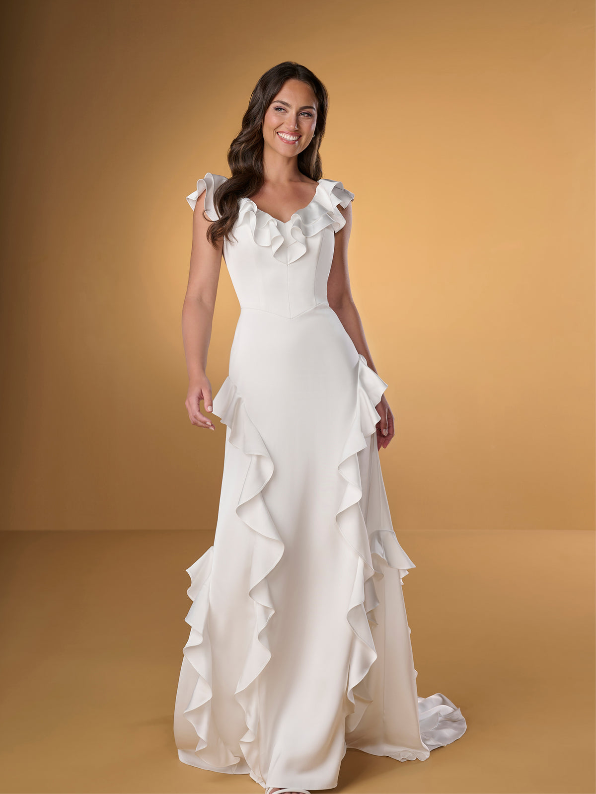 A woman modeling a white sleeveless wedding gown with ruffled cap sleeves, a fitted bodice, and cascading ruffle trim down the skirt, posed against a warm tan studio backdrop. / Slim A-line style in charmeuse with ruffles decorating V-neck and skirt.