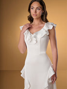 A woman modeling a white sleeveless dress with dramatic ruffle details on the shoulders and bodice, photographed against a warm tan studio background. / Slim A-line style in charmeuse with ruffles decorating V-neck and skirt.
