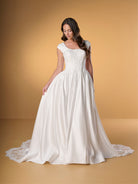 A woman modeling a white wedding gown with a full satin skirt, lace bodice, and short cap sleeves against a warm tan studio backdrop. / Ball gown with lace bodice and satin skirt. Rounded neckline with cut sleeves. Zipper back closure with buttons all the way down to hemline. Sheer cut out feature with lace motifs on train area.