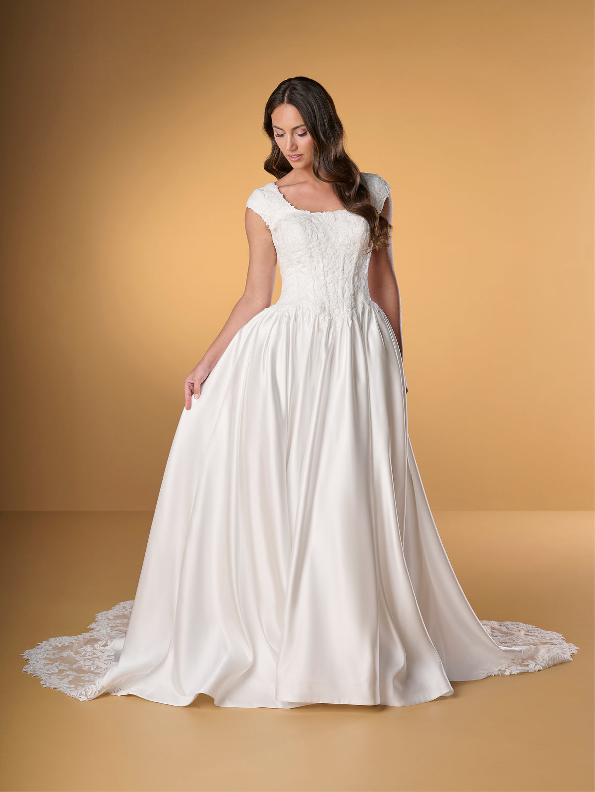 A woman modeling a white wedding gown with a full satin skirt, lace bodice, and short cap sleeves against a warm tan studio backdrop. / Ball gown with lace bodice and satin skirt. Rounded neckline with cut sleeves. Zipper back closure with buttons all the way down to hemline. Sheer cut out feature with lace motifs on train area.