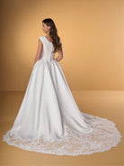 A woman modeling a white wedding gown from the back, showing a structured lace bodice, full satin skirt, and long lace-trimmed train against a warm beige studio backdrop. / Ball gown with lace bodice and satin skirt. Rounded neckline with cut sleeves. Zipper back closure with buttons all the way down to hemline. Sheer cut out feature with lace motifs on train area.