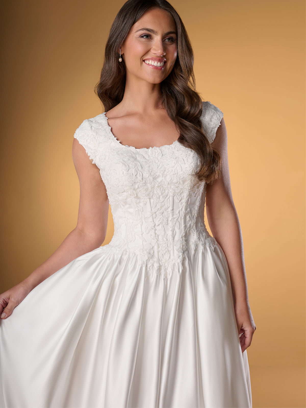 A woman modeling a white lace and satin A-line wedding dress against a warm tan studio background. / Ball gown with lace bodice and satin skirt. Rounded neckline with cut sleeves. Zipper back closure with buttons all the way down to hemline. Sheer cut out feature with lace motifs on train area.