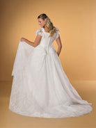 Woman wearing a white lace wedding dress against a beige background / Lace ball gown with square neckline, lace peplums and flatter sleeves. Covered buttons along zipper back closure.