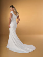 A woman modeling a white one-shoulder wedding gown with a long train, photographed against a warm tan studio backdrop. / Slim crepe style with square neckline and cup sleeves, decorated with 3D motifs in soft crinkle organza fabric. Covered buttons along zipper closure at the back.