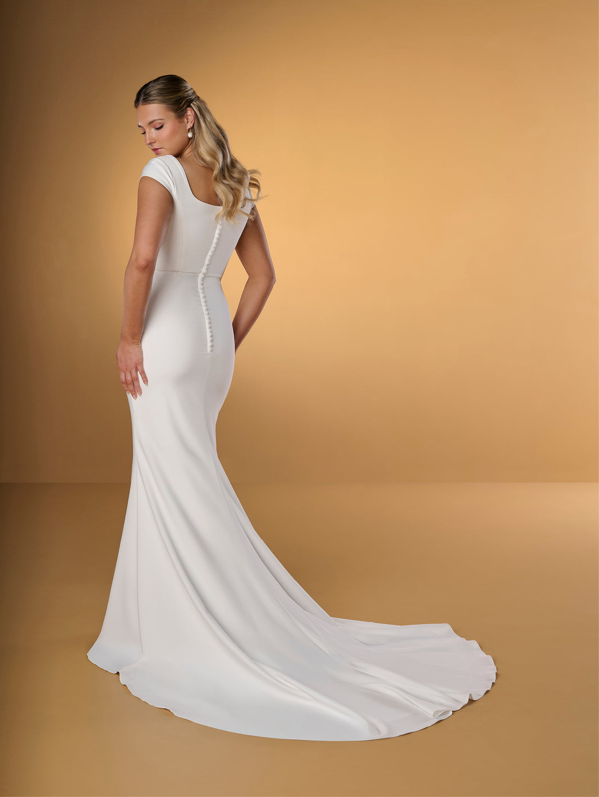 A woman modeling a white one-shoulder wedding gown with a long train, photographed against a warm tan studio backdrop. / Slim crepe style with square neckline and cup sleeves, decorated with 3D motifs in soft crinkle organza fabric. Covered buttons along zipper closure at the back.