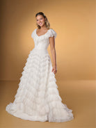 Woman wearing a white wedding dress with ruffled details against a gold background / A-line style in delicate lace with ruffles around scooped neckline and all over the skirt. Invisible zipper closure at the back.