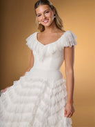 Woman wearing a white wedding dress with ruffled sleeves against a beige background / A-line style in delicate lace with ruffles around scooped neckline and all over the skirt. Invisible zipper closure at the back.