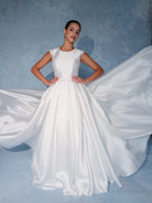 A woman modeling a white satin ball gown with a high neckline, cap sleeves, a defined waist, and a dramatic flowing train against a textured blue backdrop. / Mikado full A-line gown with scooped neckline and cup sleeves finished with narrow waist trim. Zipper/buttons back closure.