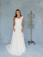 A woman modeling a sleeveless white wedding gown with a fitted bodice and flowing skirt, standing beside a vintage-style metal dress form against a textured light blue studio backdrop. / A-line style - mock 2 piece- with lace bodice and chiffon skirt. Scooped neckline, zipper/buttons back closure.
