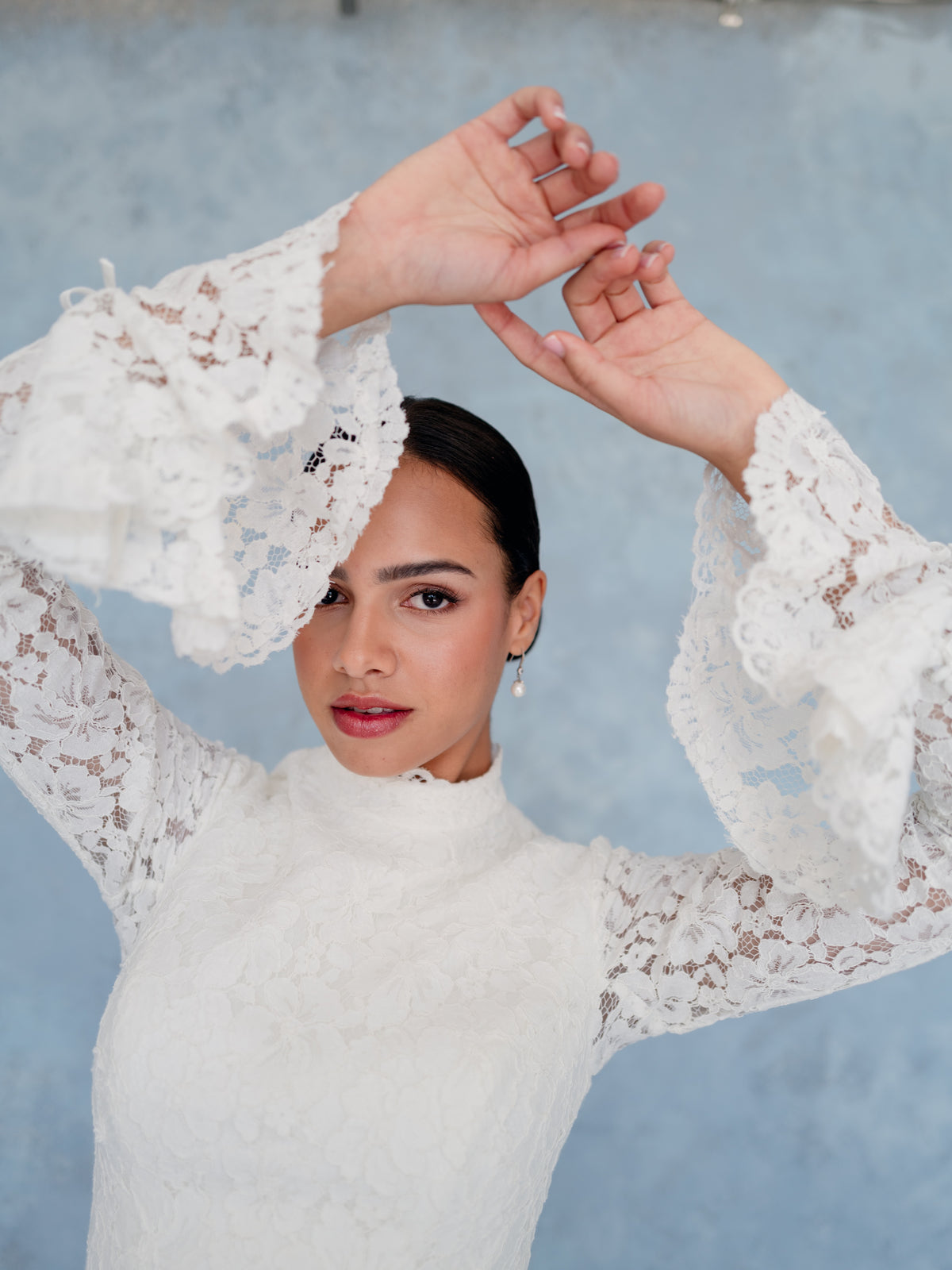 A woman wearing a white lace long-sleeve mock-neck top, posing with her hands near her head against a textured light blue background. / Slim lace style with high neck and long sheer lace sleeves. Zipper/buttons back closure - buttons all the way down to hemline.