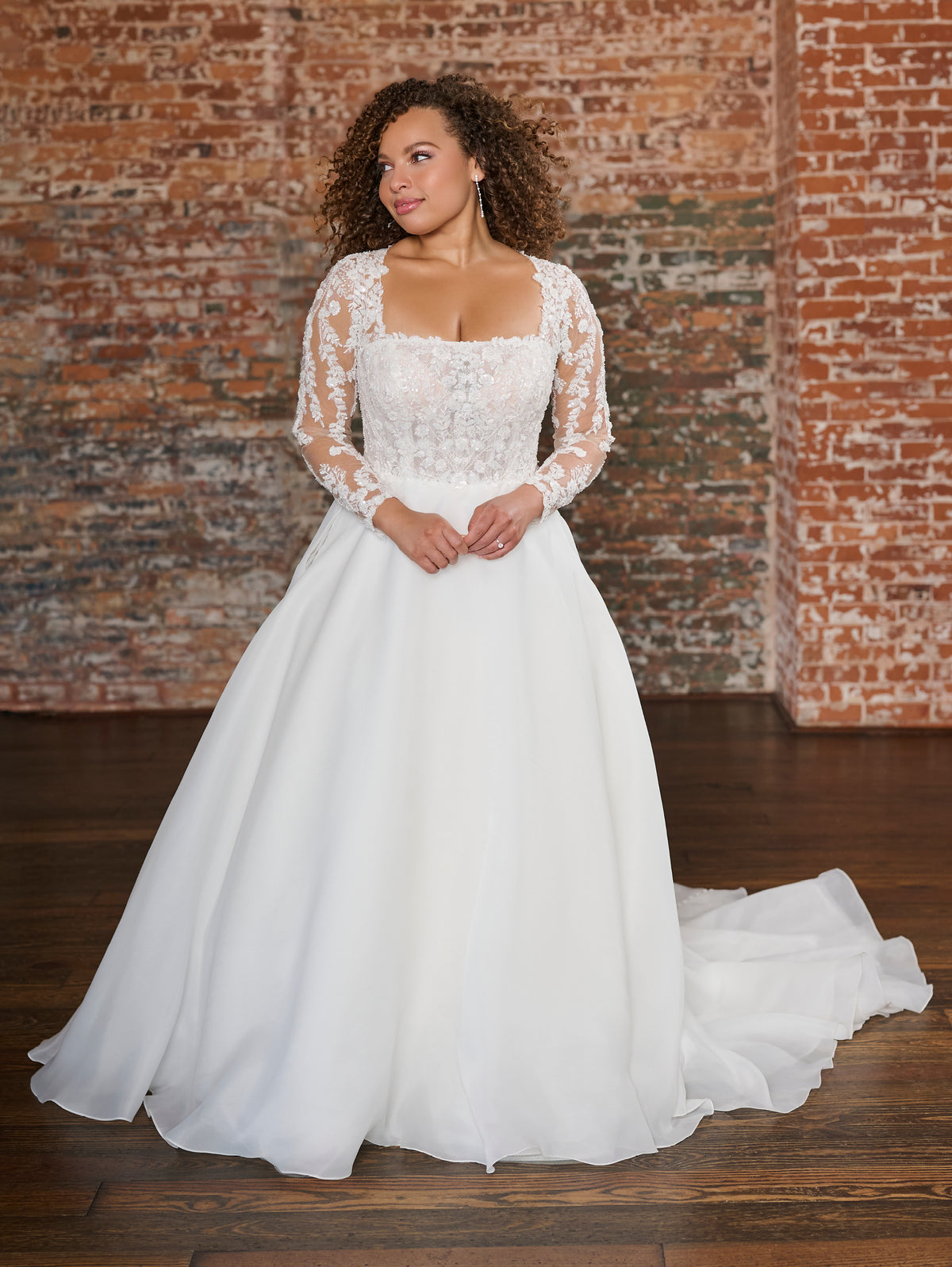 A woman wearing a white lace and tulle wedding gown stands in front of a rustic brick wall and dark wood floor. The dress has long sheer lace sleeves, a square neckline, and a full flowing skirt with a train. / This beautiful, A-line ballgown has a semi sheer bodice that is made of layers of beaded tulle, lace and plain tulle to create the perfect amount of coverage. Boning has been placed to perfectly support the body through the bodice. The flattering square neckline is complimented by long, sheer sleeves