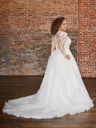 A woman wearing a white wedding dress with lace long sleeves and a full tulle skirt, standing in a room with a brick wall and wooden floor. / This beautiful, A-line ballgown has a semi sheer bodice that is made of layers of beaded tulle, lace and plain tulle to create the perfect amount of coverage. Boning has been placed to perfectly support the body through the bodice. The flattering square neckline is complimented by long, sheer sleeves and an illusion back bodice. The skirt begins at the natural