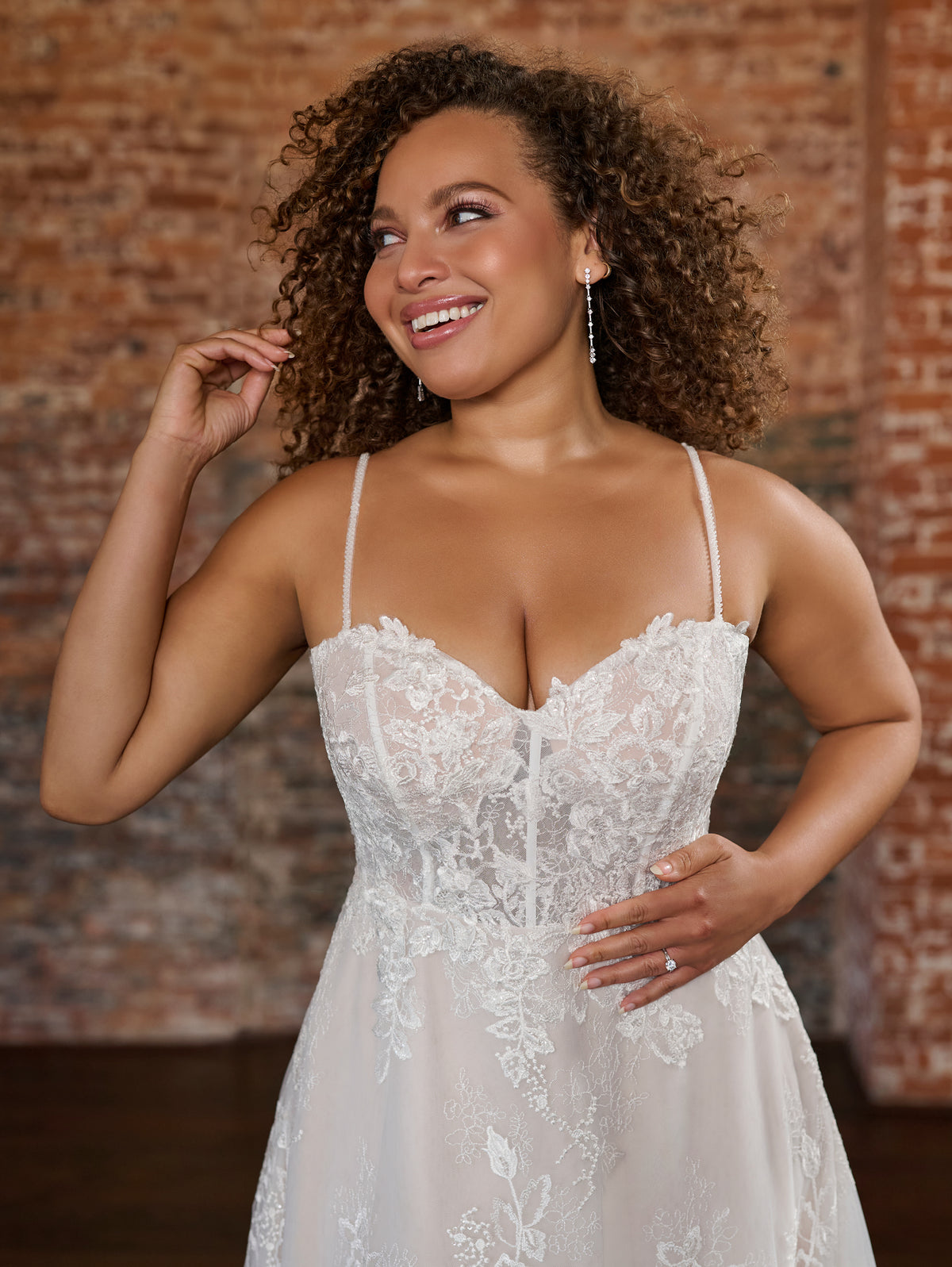 A woman wearing a white lace wedding dress with thin spaghetti straps and a sweetheart neckline, posing in front of a rustic brick wall background. / Soft romantic A-line dress in airy all over lace with sweetheart neckline and short balloon sleeves. Semi sheer bodice decorated with beautiful floral lace. Covered buttons at the back closure, long train.