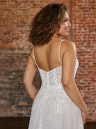 A woman wearing a white lace bridal gown is shown from the back, standing in front of a rustic brick wall. The dress has thin spaghetti straps, a fitted lace bodice, and a flowing skirt. / Soft romantic A-line dress in airy all over lace with sweetheart neckline and short balloon sleeves. Semi sheer bodice decorated with beautiful floral lace. Covered buttons at the back closure, long train.