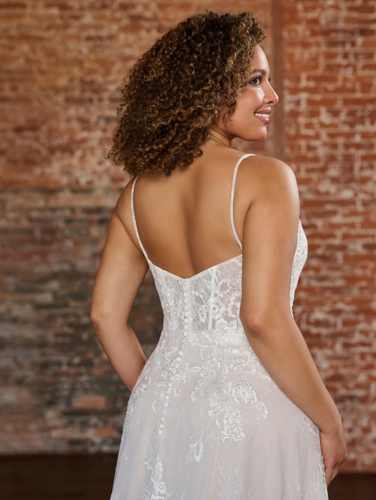 A woman wearing a white lace bridal gown is shown from the back, standing in front of a rustic brick wall. The dress has thin spaghetti straps, a fitted lace bodice, and a flowing skirt. / Soft romantic A-line dress in airy all over lace with sweetheart neckline and short balloon sleeves. Semi sheer bodice decorated with beautiful floral lace. Covered buttons at the back closure, long train.
