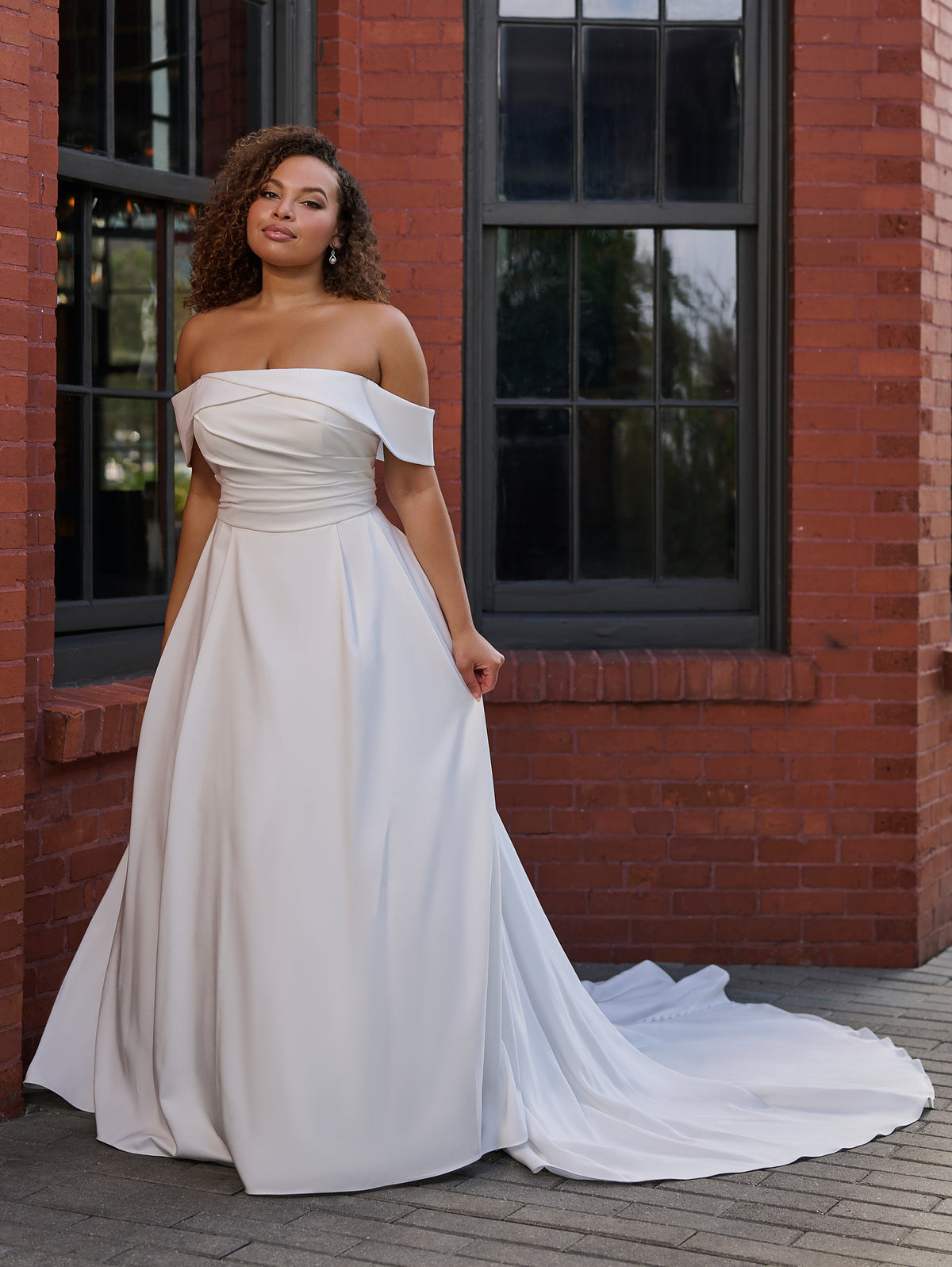 Woman wearing a white off-shoulder gown standing in front of a brick building. / Clean satin A-line style with draped bodice and off-shoulder straps. Covered buttons all the way to hemline on CB closure finishing the look.