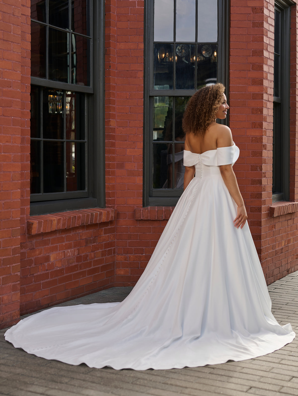 A woman wearing a white off-the-shoulder ball gown with a long train, standing outdoors in front of a red brick building with dark-framed windows. / Clean satin A-line style with draped bodice and off-shoulder straps. Covered buttons all the way to hemline on CB closure finishing the look.