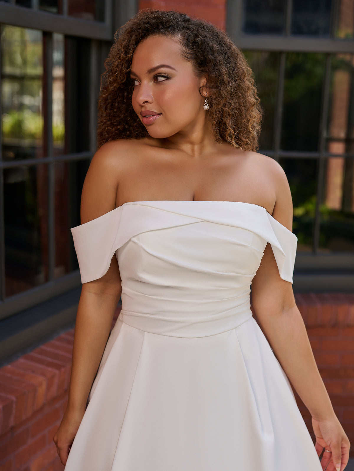 A woman wearing an off-the-shoulder white or ivory formal dress with a structured bodice and flared skirt, standing outdoors in front of a brick building with large windows. / Clean satin A-line style with draped bodice and off-shoulder straps. Covered buttons all the way to hemline on CB closure finishing the look.