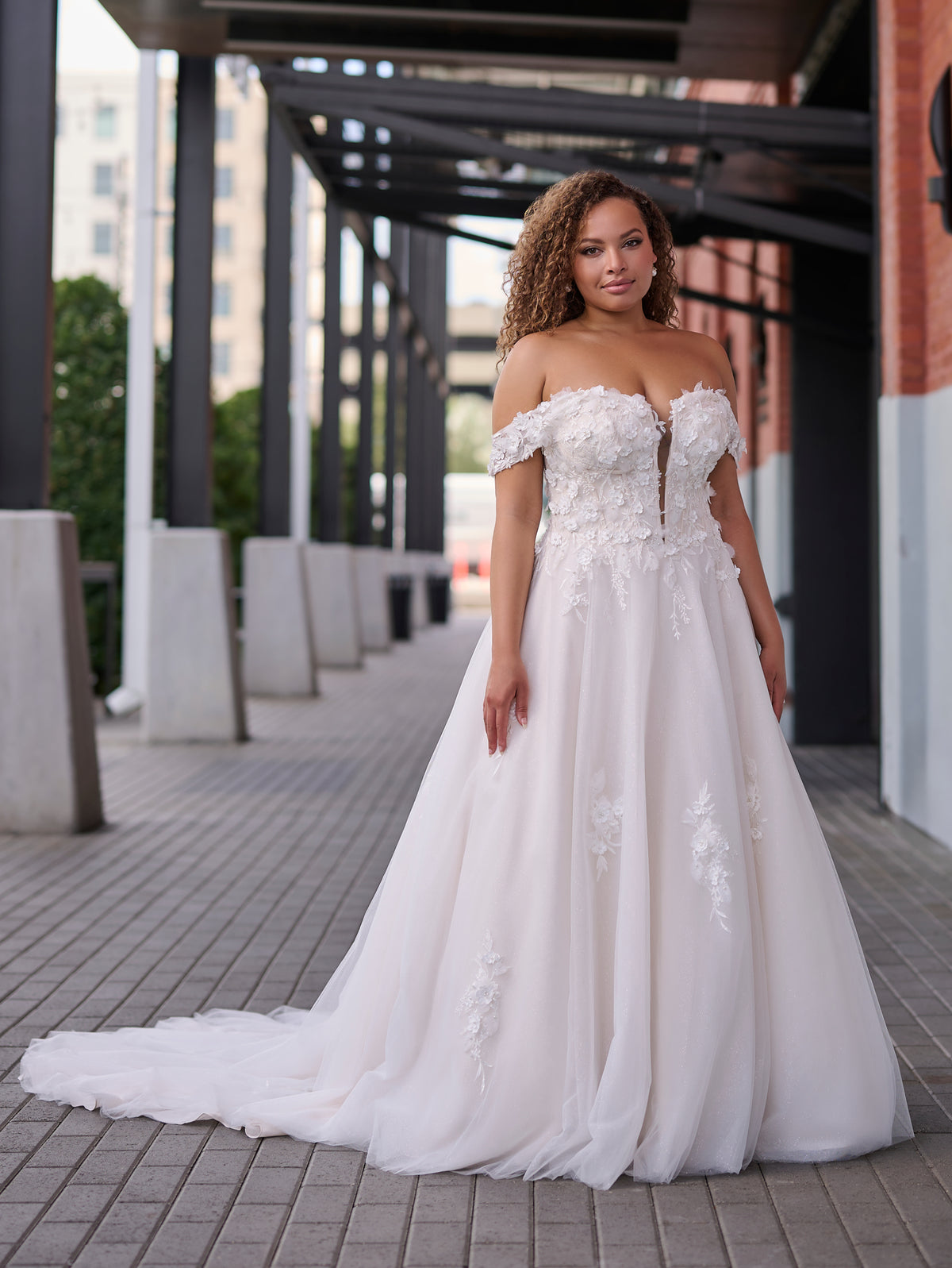 A woman wearing an off-the-shoulder A-line wedding gown with a sweetheart neckline, lace applique bodice, and a long train, standing in an outdoor covered walkway. / Soft A-line gown in tulle with 3D lace motifs decorating bodice with sweetheart neckline and deep illusion v on center front. Off-shoulder straps. Long train.