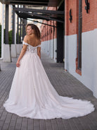 A woman wearing a voluminous off-the-shoulder white wedding gown with floral lace applique and a long train, photographed outdoors on a paved walkway beside a brick and white building. / Soft A-line gown in tulle with 3D lace motifs decorating bodice with sweetheart neckline and deep illusion v on center front. Off-shoulder straps. Long train.