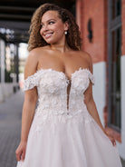 A woman wearing a strapless white wedding gown with floral lace applique and a tulle skirt, photographed outdoors in front of a brick building. / Soft A-line gown in tulle with 3D lace motifs decorating bodice with sweetheart neckline and deep illusion v on center front. Off-shoulder straps. Long train.