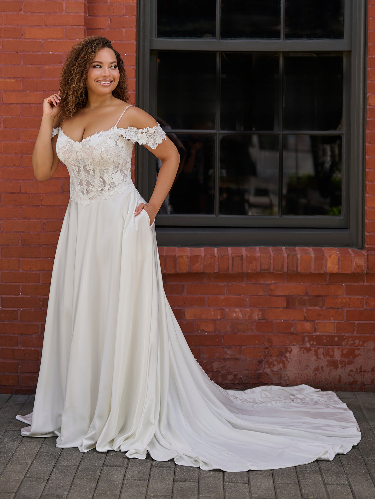 A woman wearing an off-the-shoulder A-line wedding dress with a lace bodice and flowing satin skirt, standing in front of a red brick wall with a dark window. / Off the shoulder lace and charmeuse soft A- line gown with sheer illusion bodice.