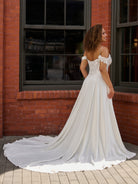 A woman wearing a white off-the-shoulder wedding gown with a long train, photographed outdoors in front of a red brick building with dark-framed windows. / Off the shoulder lace and charmeuse soft A- line gown with sheer illusion bodice.