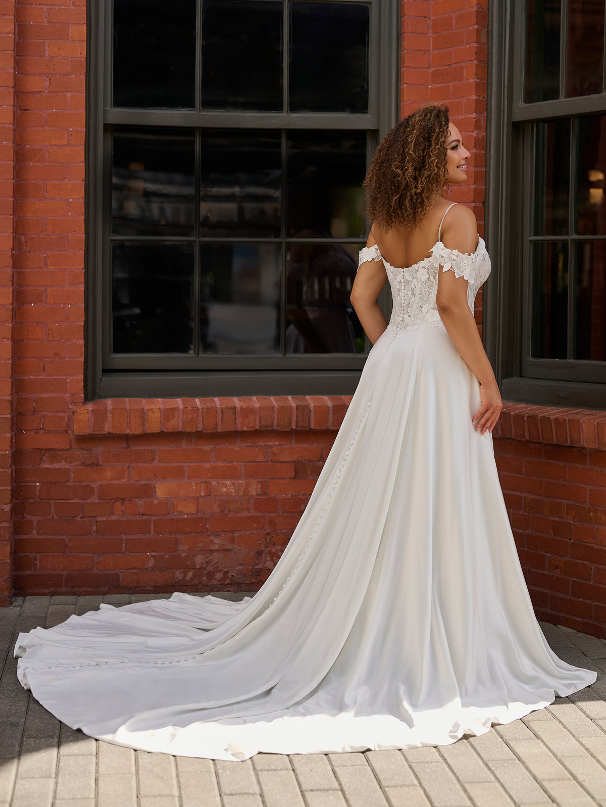 A woman wearing a white off-the-shoulder wedding gown with a long train, photographed outdoors in front of a red brick building with dark-framed windows. / Off the shoulder lace and charmeuse soft A- line gown with sheer illusion bodice.
