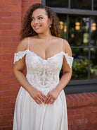 A woman wearing a white lace off-the-shoulder wedding dress stands outdoors against a red brick wall and dark window background. / Off the shoulder lace and charmeuse soft A- line gown with sheer illusion bodice.