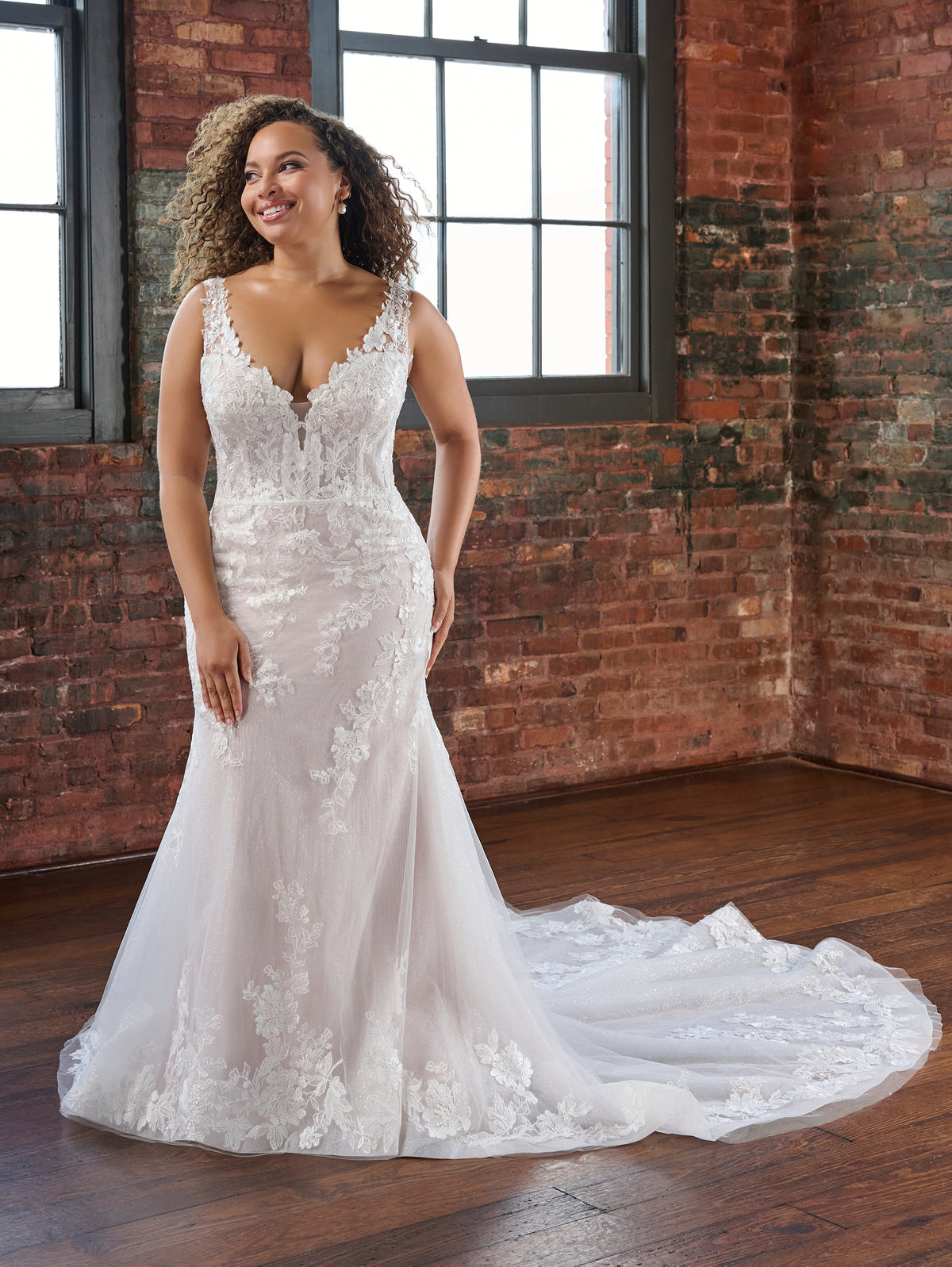 A woman models a white lace mermaid-style wedding gown with a deep V neckline, fitted bodice, and long train in an indoor room with exposed brick walls and wood floors. / This slim and sexy gown has placed lace appliques that sit on a layer of tulle that is placed over sparkle tulle. The semi sheer bodice leads to sheer straps and a sheer back bodice. There are flattering pleats at the back and a long sheer train.
