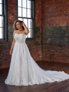 A woman modeling a strapless white wedding gown with a fitted lace bodice and a long sheer tulle train, standing in a brick warehouse room with large windows. / Soft A-line style with square neckline, this style features floral lace motifs with 3D petals.
