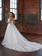 A woman wearing a white lace wedding gown with a long train stands in a brick warehouse-style room with large windows and dark wood floors. / Soft A-line style with square neckline, this style features floral lace motifs with 3D petals.