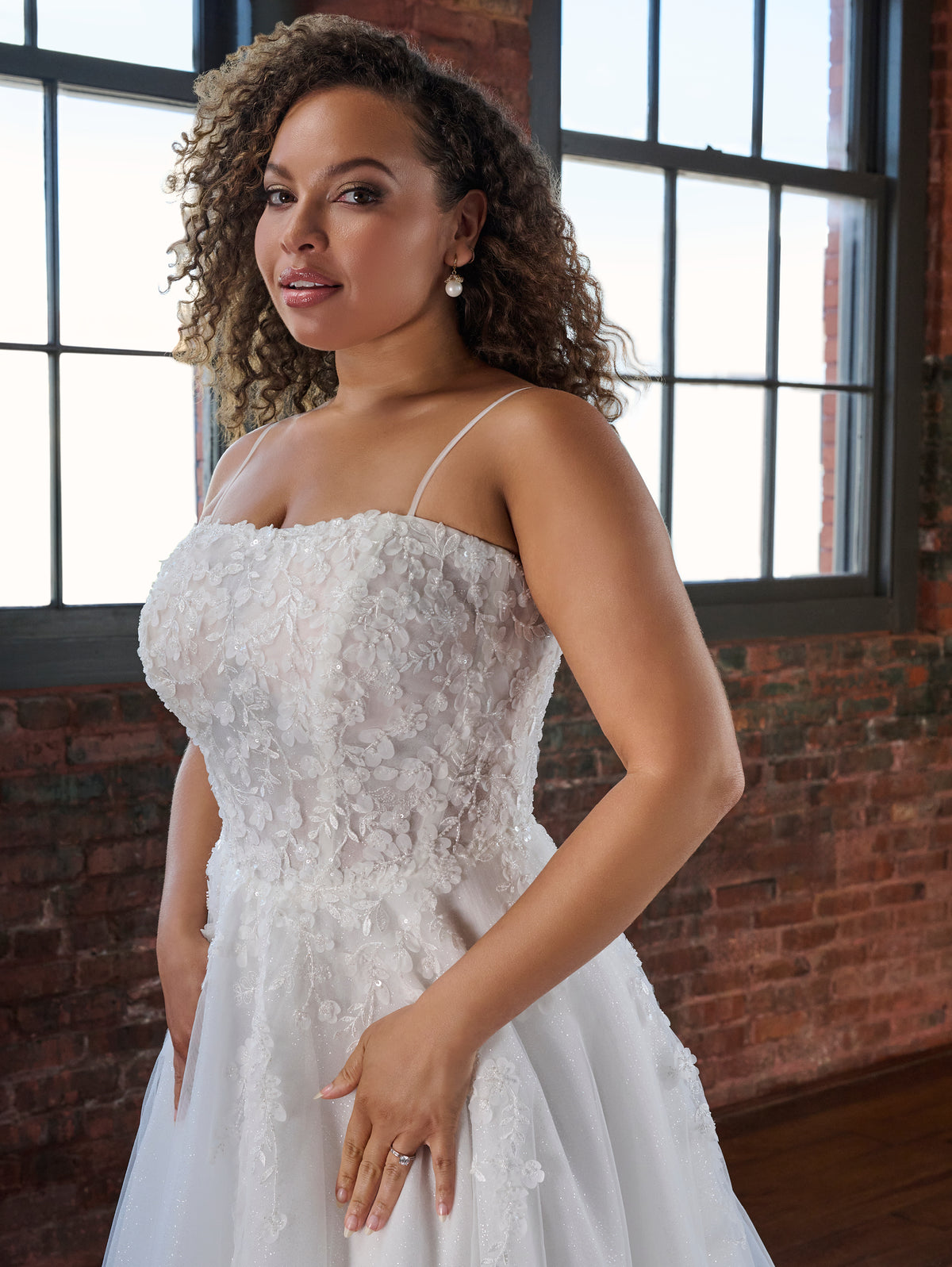 A woman wearing a white strapless lace wedding gown stands indoors in front of large windows and a brick wall. The dress has a fitted lace bodice, thin spaghetti straps, and a full tulle skirt. / Soft A-line style with square neckline, this style features floral lace motifs with 3D petals.
