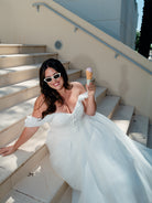 A woman in a white off-the-shoulder wedding gown and white cat-eye sunglasses sits on outdoor stone stairs, holding an ice cream cone. / Lace bodice with tulle draping and detachable off the shoulder swags and a full, organza skirt.