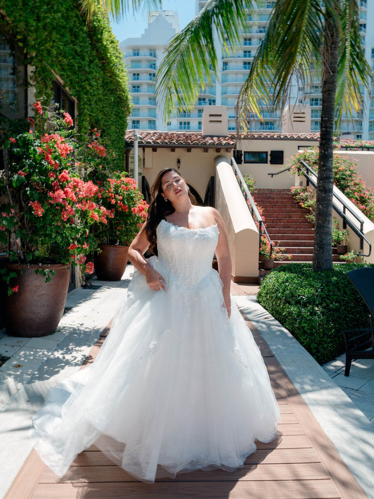 A woman wearing a strapless white tulle wedding gown stands on a wooden walkway outdoors, with palm trees, potted flowers, stairs, and a building visible in the background. / Basque waist all over lace and lace applique ballgown.