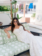 A woman reclining on a patterned outdoor sofa wearing a strapless white lace wedding gown and sunglasses in a bright patio setting. / Lace A-line style with sweetheart neckline and lace up back closure.