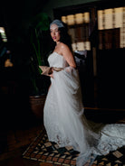A woman in a strapless white lace wedding gown with a sheer veil and a long flowing train stands in a dimly lit interior. She holds a small tan clutch, and the background includes dark furniture and warm-toned wall panels. / Placed lace slim gown with scallop detail.