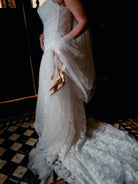 A bride in a white lace wedding gown with a long tulle train, holding a pair of nude pointed-toe heels, photographed indoors against a dark background. / Placed lace slim gown with scallop detail.