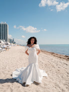 A woman wearing a white off-the-shoulder mermaid-style wedding gown stands on a sandy beach with the ocean and high-rise buildings in the background. / Mermaid style in double face satin with scooped neckline and attached dramatic off-shoulder straps. Bodice decorated with beaded motifs with small 3D flowers. Zipper/buttons back closure.
