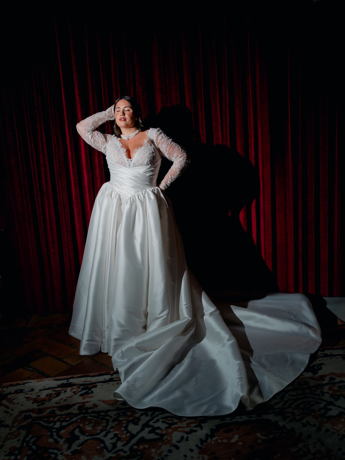 A woman wearing a white wedding gown with long sheer lace sleeves and a full satin skirt, standing in front of a dark red curtain and casting a long shadow. / Ball gown with lace bodice and sleeves, and mikado skirt. Pleated cummerbund on waist. Zipper/buttons back closure.